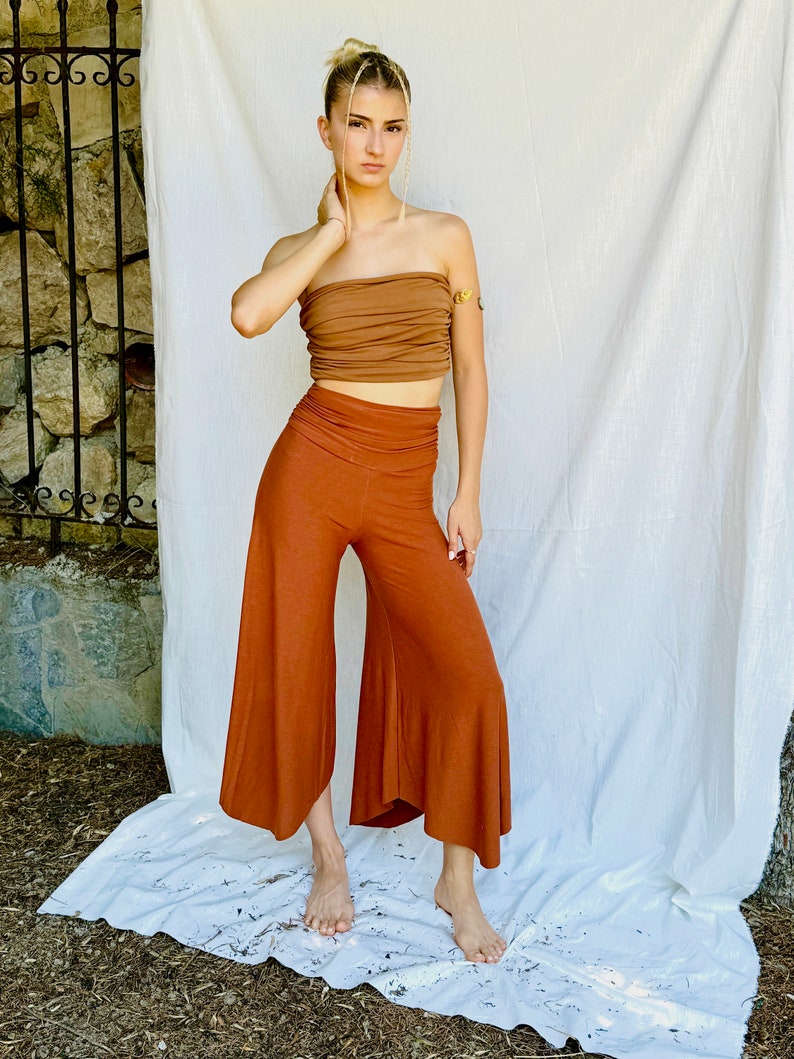 a young woman with blonde hair styled in a high bun is modeling a terracottacolored outfit.  she's wearing highwaisted, wideleg culottes that flare out slightly at the hem. the pants are a relaxed fit, and the fabric appears soft and lightweight.  her top is a matching burnt orange bandeau or tube top that is slightly gathered. she's barefoot and stands on a white cloth backdrop, possibly outdoors against a stone wall and sheer white fabric.  the overall style is bohemian and relaxed.