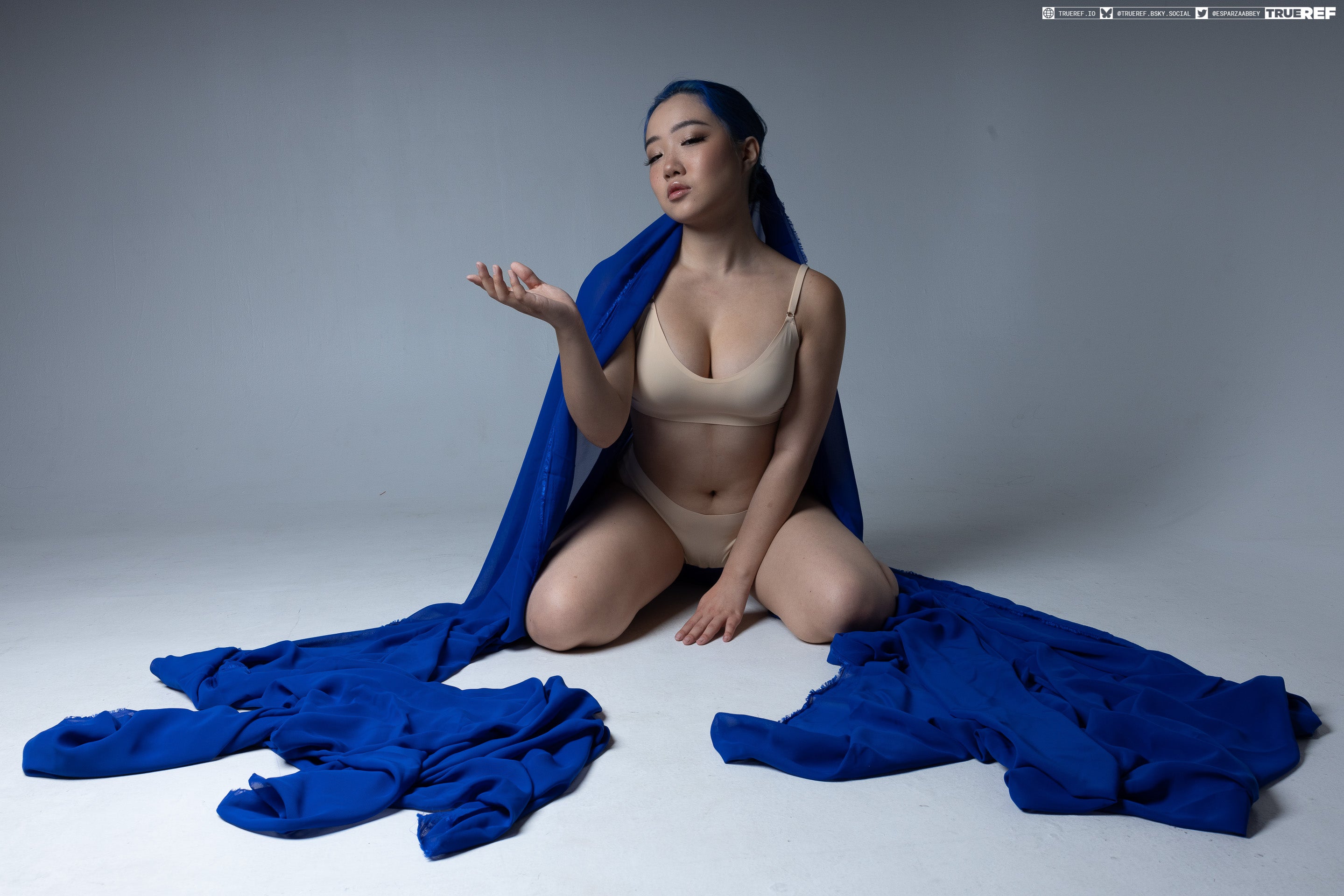 a woman with blue hair kneels on a white surface. she is wearing underwear and a bra and is draped in a blue fabric. her right arm is outstretched with her hand open and facing up.