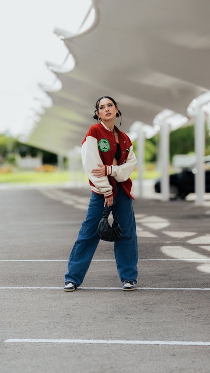 a stylish young woman in a red and white varsity jacket, blue jeans, and black sneakers stands confidently in a parking lot, looking up. she carries a black handbag. the background features a modern architectural structure with a white canopy. the overall style is casual yet trendy, with a focus on streetwear fashion.