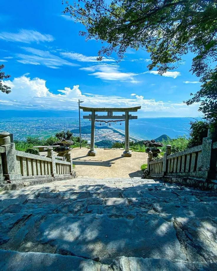 the image shows a scenic view from the top of a stone staircase, leading to a traditional japanese torii gate. beyond the gate, a cityscape stretches towards the sea, with a clear blue sky and scattered clouds. the composition is framed by trees and stone railings, creating a sense of depth and perspective.