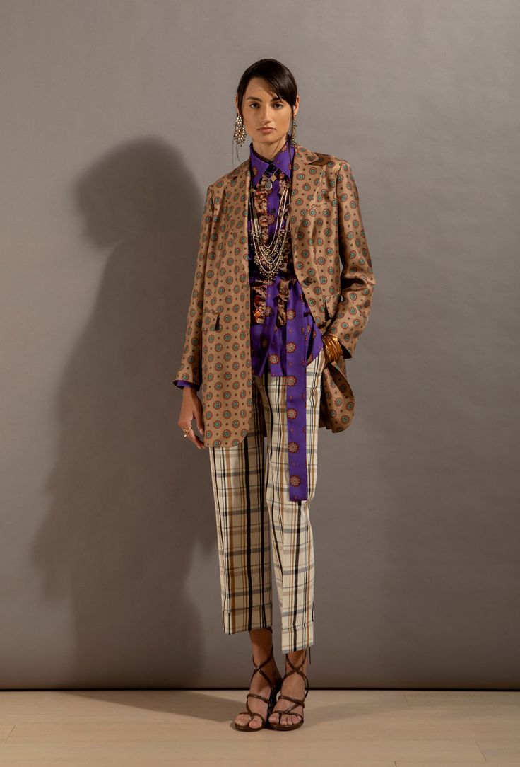 a fullbody shot of a fashion model against a muted grey backdrop. the model is wearing a tan patterned blazer over a purple patterned blouse. she's also wearing cream and brown plaid cropped trousers, brown heeled sandals, and an array of jewelry including a layered necklace and earrings. the overall style is sophisticated and slightly bohemian. the image is professionally lit and styled for a fashion catalog or editorial.