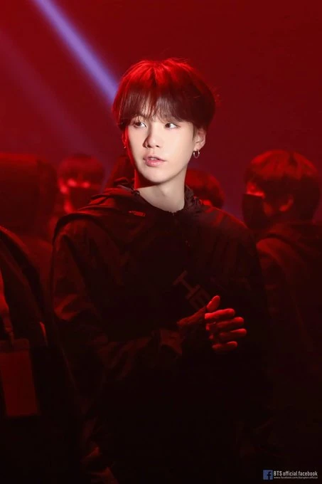 this image features suga from the south korean boy band bts standing on a stage. he is dressed in a black hoodie and illuminated by a red spotlight. he has short, dark hair styled in a bowl cut and is wearing hoop earrings. in the background, there is a blurred audience, and the overall scene suggests a performance or concert setting.