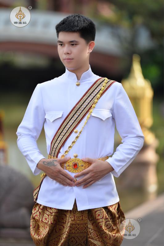 a thai man is dressed in a traditional thai outfit for a wedding ceremony. he's wearing a white longsleeve shirt with a red sash, a gold belt and gold patterned pants.