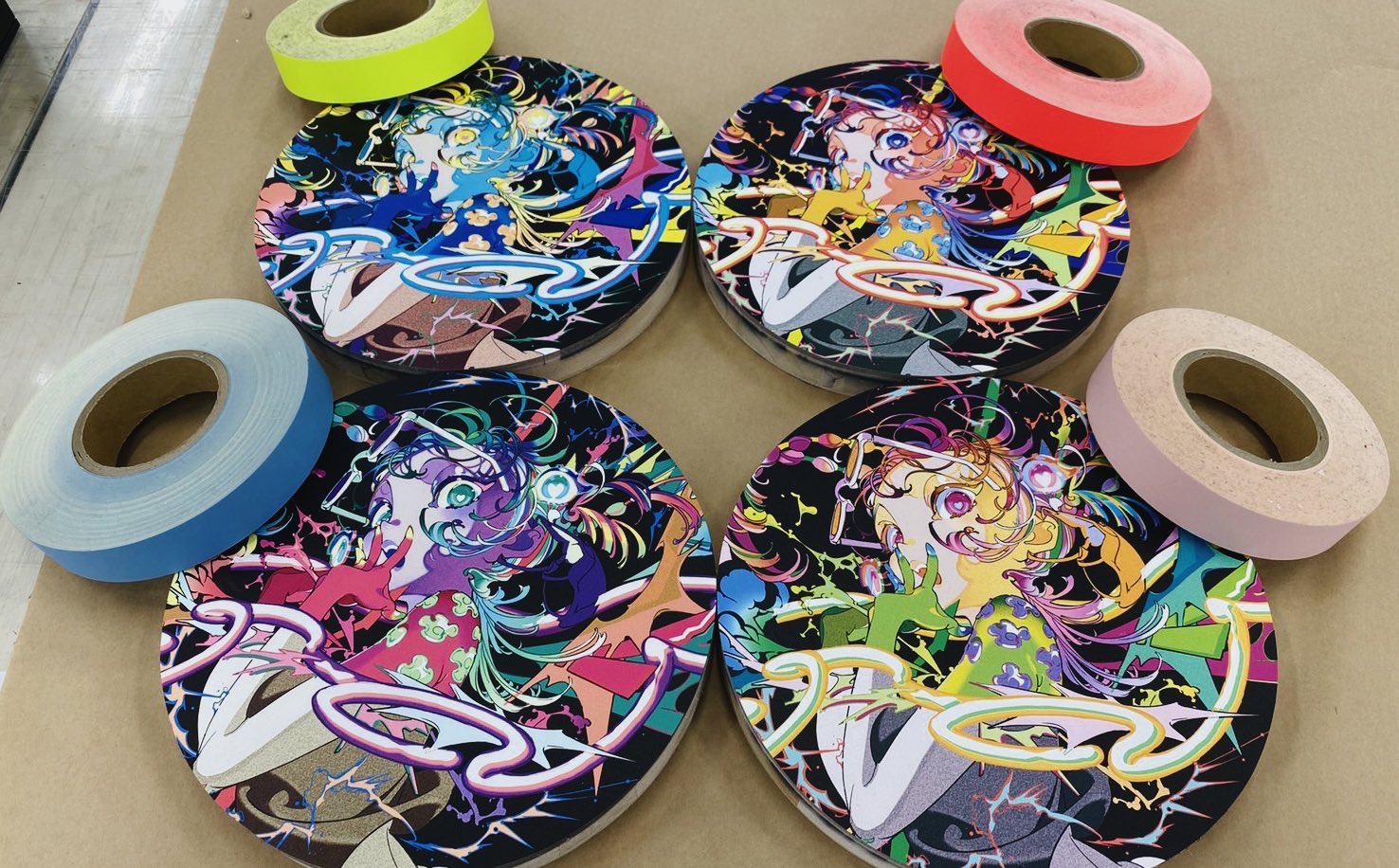 the image shows four vibrant art prints arranged in a circular pattern. each print features the same animestyle female character with multicolored hair and clothing set against a black background with neon accents. surrounding the prints are five rolls of colorful tape in fluorescent yellow, pink, blue, beige, and a lighter shade of pink. the presence of the tape suggests that these prints are being prepared for shipping or display.  the overall aesthetic is bright, playful, and likely inspired by japanese pop culture.