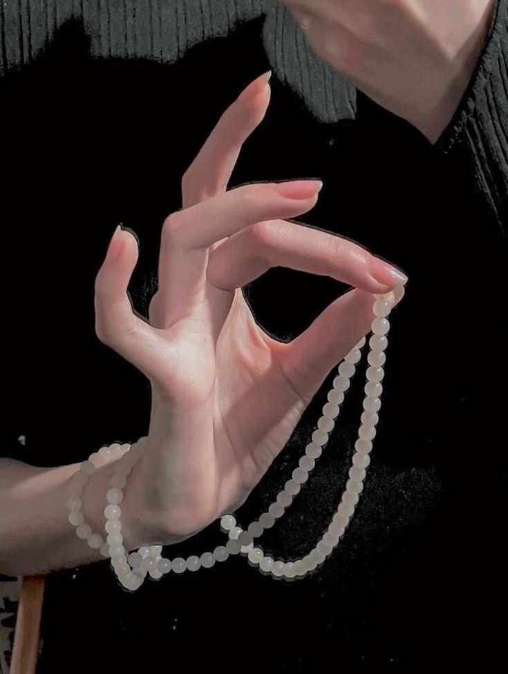 the image shows a hand holding a string of white beads. the hand has long, manicured nails and pale skin. the person is wearing a black sweater. the background is dark.
