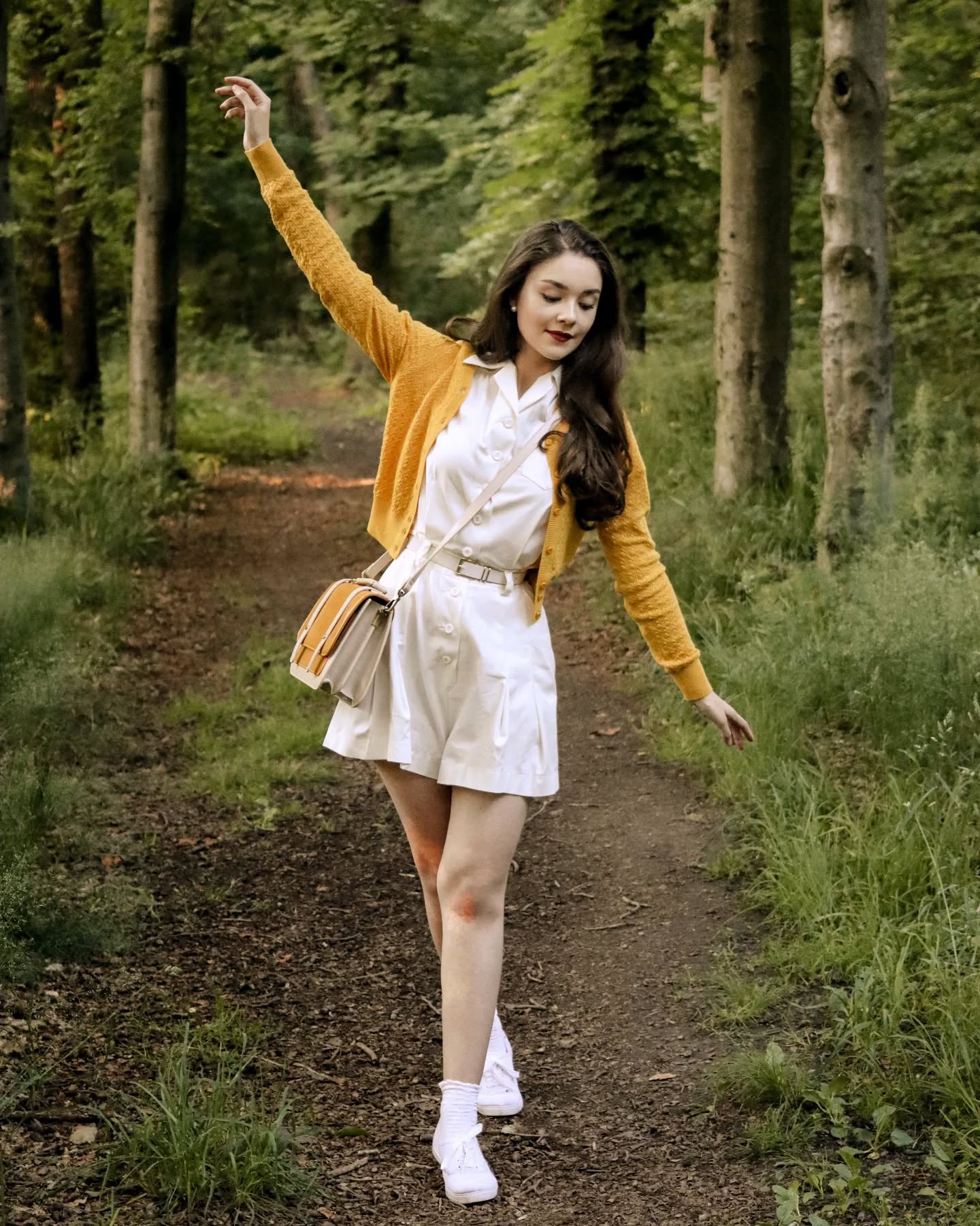 a young woman in a stylish outfit strolls through a sundappled forest path. she's wearing a creamcolored pleated dress, a mustardyellow cardigan, and white sneakers. a light beige crossbody bag hangs from her shoulder. her brown hair is long and flowing, and she has a joyful expression on her face. the scene evokes a sense of carefree summer days and classic vintage fashion. the forest setting provides a backdrop of lush green trees and dappled sunlight, enhancing the overall aesthetic.
