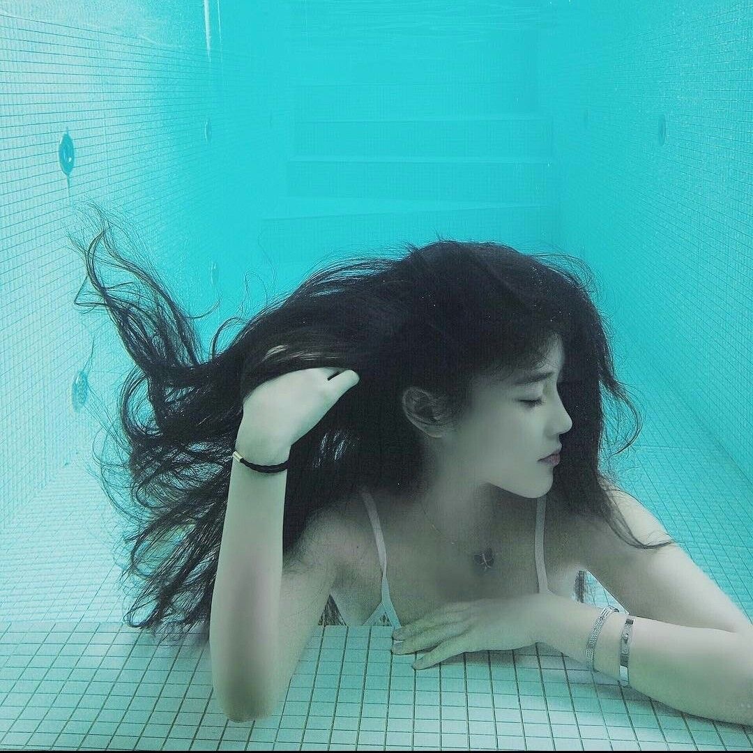 a young woman with long dark hair is submerged in a swimming pool. she is wearing a white bikini top and has her eyes closed, her hair floating around her head. the pool is tiled in a light blue and white mosaic pattern. the water is clear and a bright turquoise color. the overall feeling is one of serenity and peacefulness.
