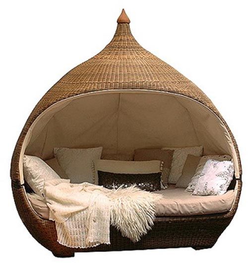 a round outdoor daybed made of wicker with a domeshaped cover is shown in a full shot. the bed has a tan cushion and several pillows, including some decorative ones. a white blanket and a fur throw are draped on the bed.