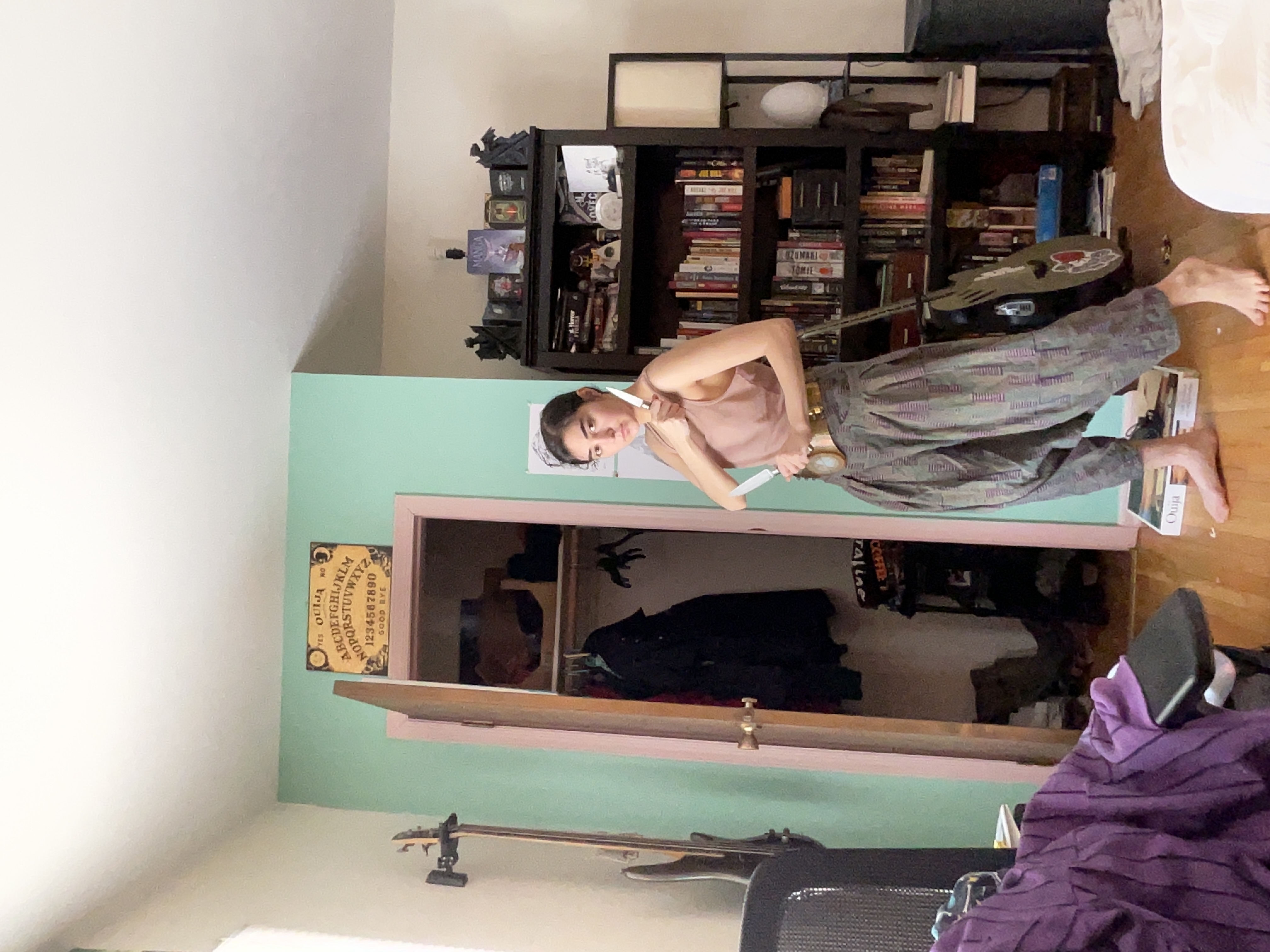 woman is standing in a room with green walls. she is wearing loosefitting camouflage pants and a light pink top. she holds a knife in her right hand. a guitar leans against a wall next to a closet.  a bookshelf is filled with books in the background, along with a ouija board hanging on the wall. a purple blanket is draped over a chair in the foreground. the room is cluttered but decorated with a bohemian feel.