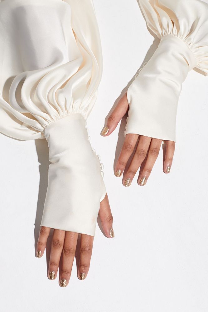 loseup image showcasing a woman's hands wearing a pair of ivory satin gloves. the gloves are attached to long puff sleeves of a matching ivory satin top or blouse. the sleeves and gloves are detailed with button cuffs. the woman's nails are elegantly manicured with a gold glitter polish. the overall aesthetic is minimalist, elegant, and sophisticated, suggestive of bridal or wedding attire. the image is welllit, with a clean white background that emphasizes the texture and color of the garment.