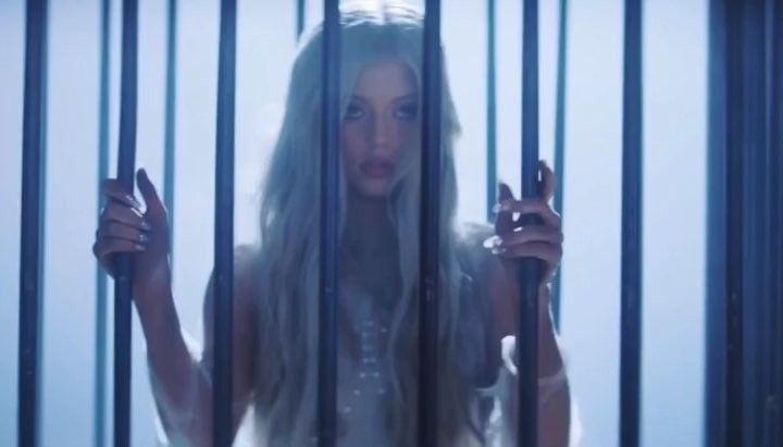 a woman with long blonde hair stands behind dark metal bars, seemingly imprisoned or held captive. her hands grip the bars, conveying a sense of longing, frustration, or vulnerability. the lighting is cool and somewhat muted, highlighting her pale skin and hair against the dark bars. the image evokes themes of imprisonment, captivity, and the desire for freedom.
