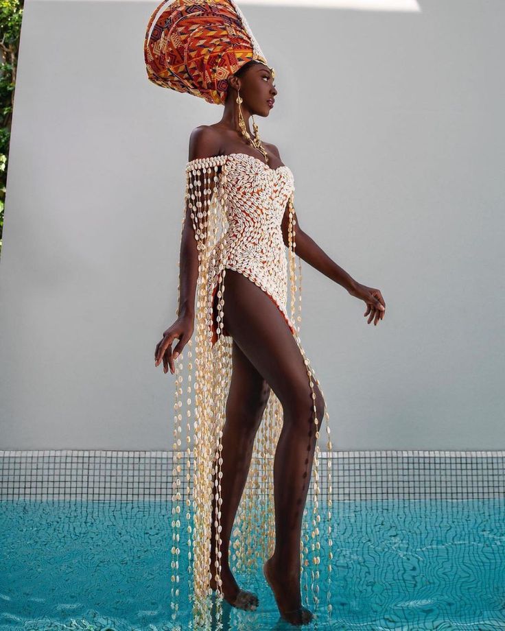 a woman is standing in a pool, wearing a dress made of cowrie shells, a large patterned head wrap, and jewelry. she is posing and looking off to the side.