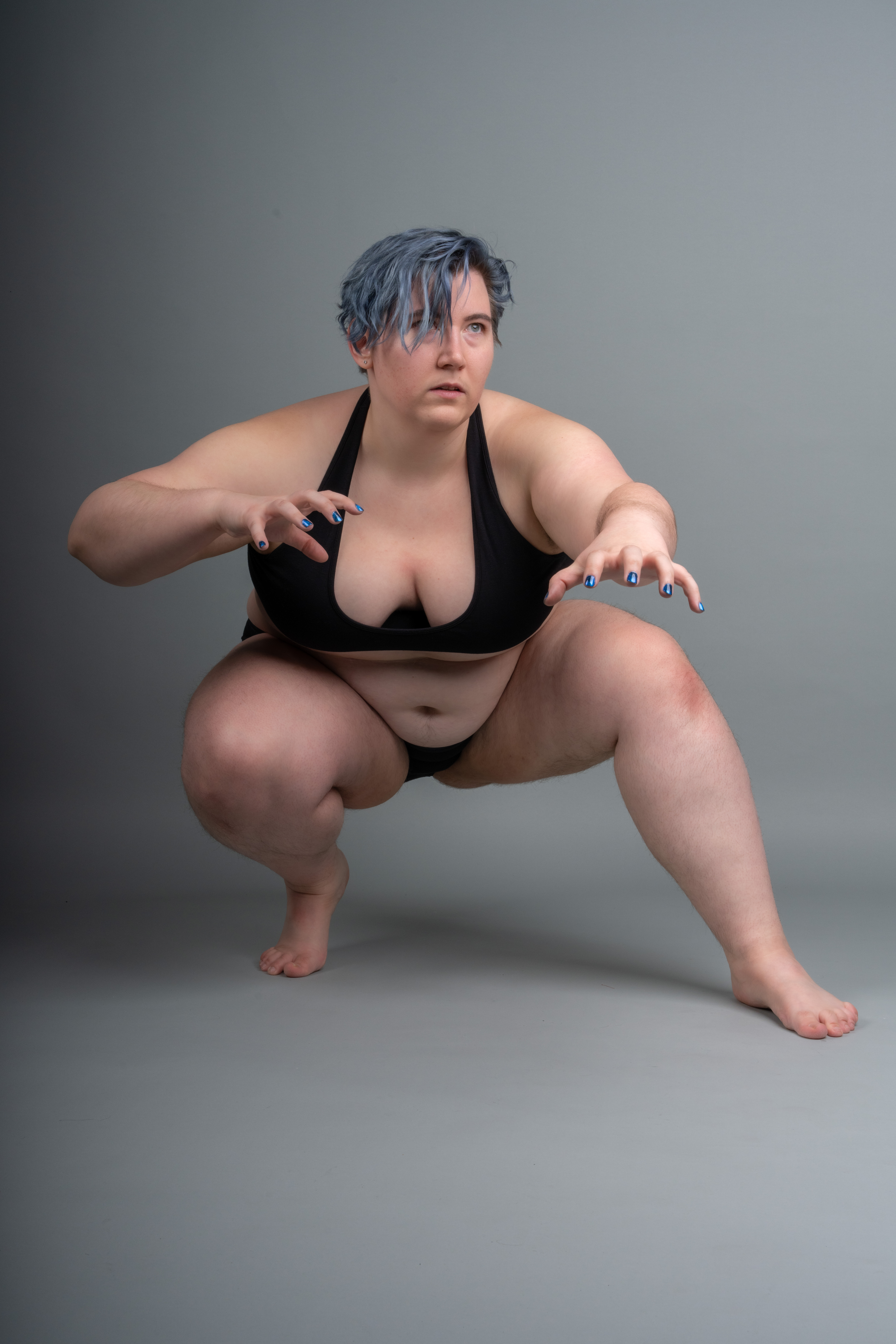 a woman with short blue hair is squatting on a gray background, wearing a black bikini and looking off to the side with a concerned expression. her hands are outstretched in front of her, fingers slightly curled.