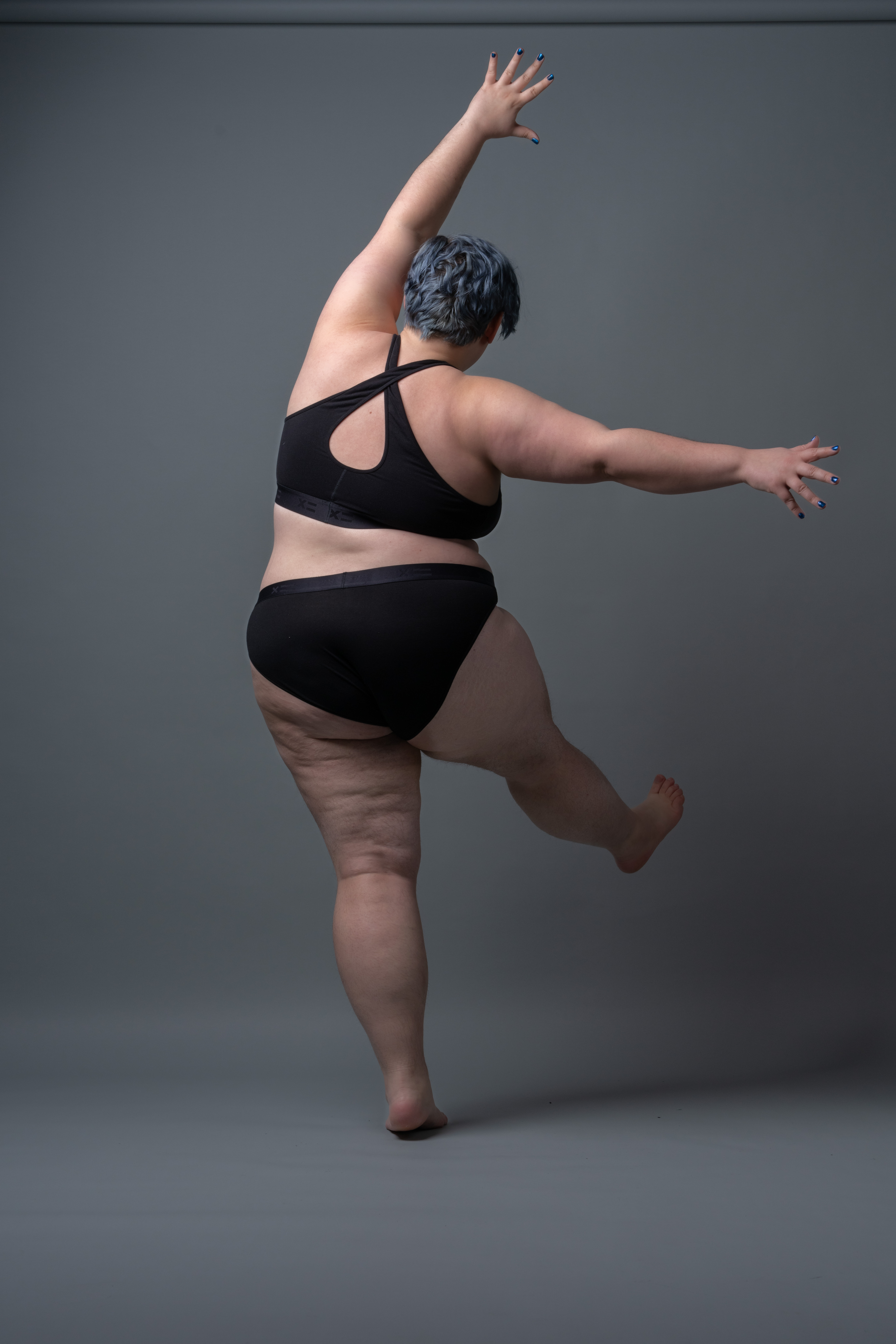 a plus size woman with short blue hair stands with her back to the viewer, wearing a black sports bra and black underwear. she stretches her body, extending one arm upward and the other out to the side. one leg is planted while the other is lifted slightly off the ground. the background is a solid gray.