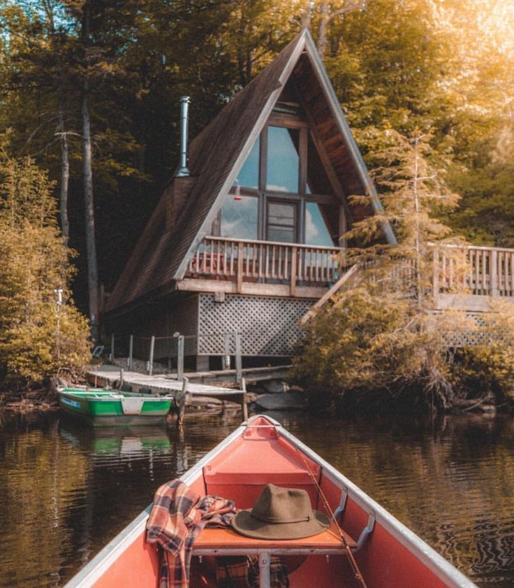 the image showcases a tranquil scene of an aframe cabin nestled amidst a lush forest bordering a calm lake.  a red canoe is in the foreground, positioned on the water, heading towards the cabin. a green canoe is docked at a small wooden pier near the cabin.  the sun creates a warm, golden light filtering through the trees, giving the scene an autumnal feel.  a plaid blanket and a hat are visible inside the red canoe. the overall atmosphere is serene and peaceful, suggesting a tranquil getaway in nature.