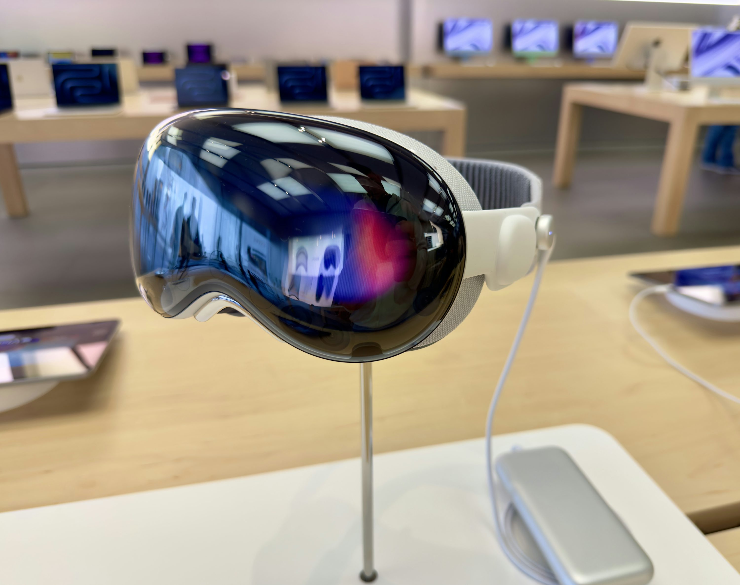 an apple vision pro vr headset is displayed on a stand in what appears to be an apple store. the headset features a dark reflective lens and a white headband. a white power cord connects the headset to a battery pack on the table. in the background, various apple products are arranged on tables.