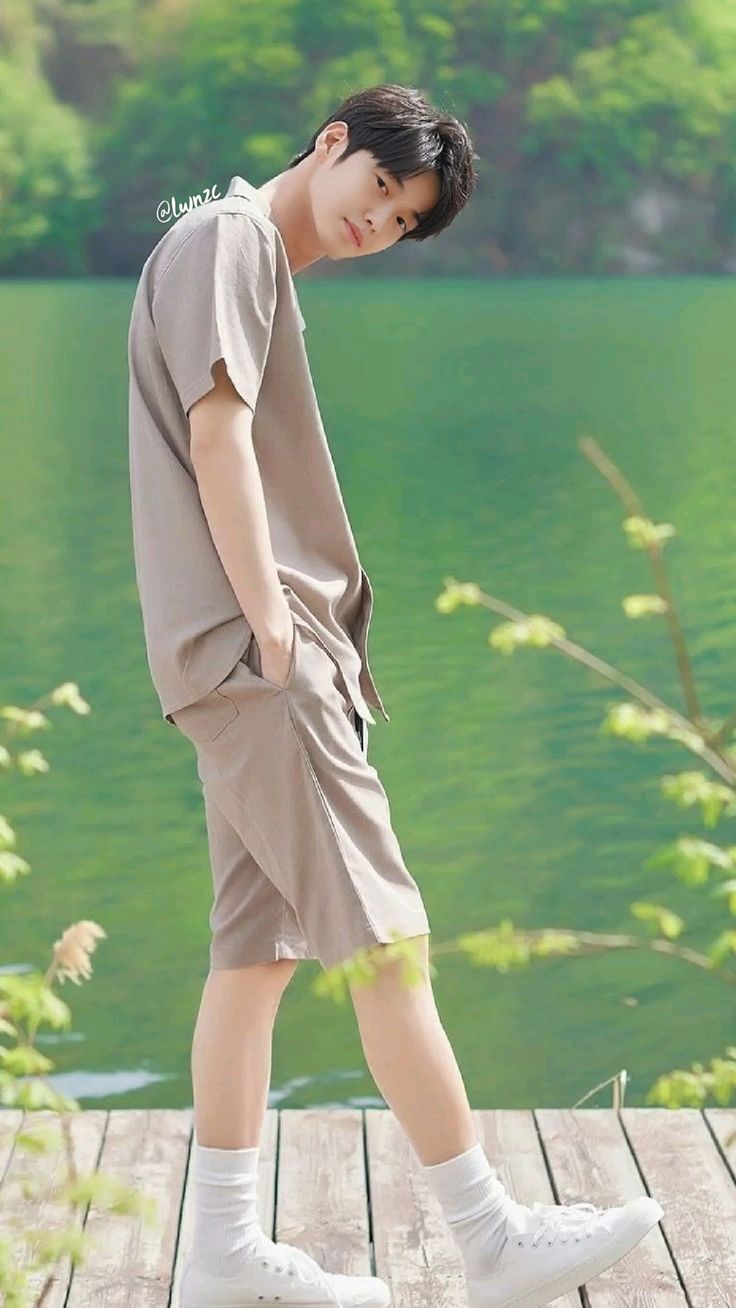 a young asian man in casual summer attire stands on a wooden dock overlooking a serene lake. he is wearing a beige buttonup shirt with matching shorts, white socks, and white sneakers. he has short black hair and a relaxed posture, with one hand casually tucked into his pocket.  the environment suggests a peaceful and sunny day, with soft, natural light illuminating the scene. the image evokes a sense of laidback style and tranquility.