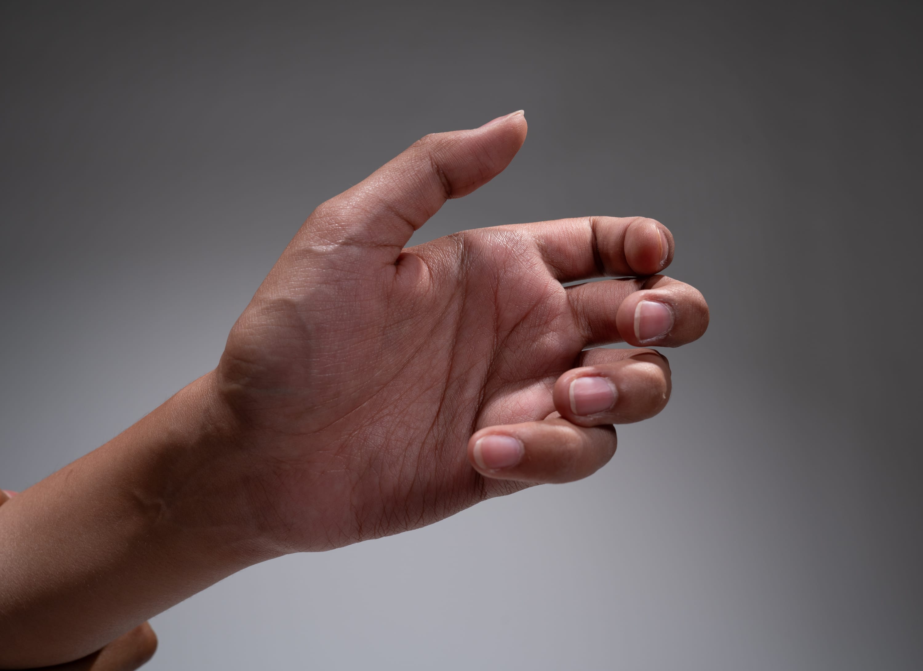 a hand is extended in front of a gray background, with the fingers slightly curled inward. the hand appears to be reaching out for something. the image is lit with soft, artificial lighting, giving the skin a warm tone.
