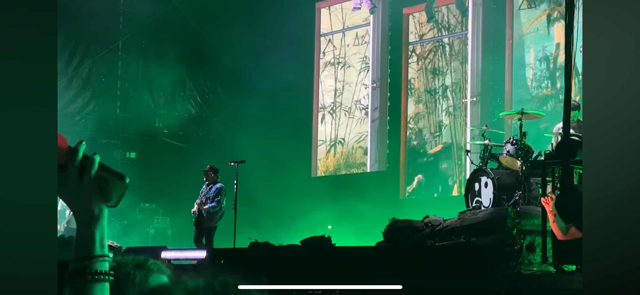 a concert stage is lit up green with a guitarist playing in the foreground, and a drummer playing in the background with a large screen behind them. the audience can be seen in the foreground, with one person's hand and phone in focus. the lights are bright and green, and the atmosphere is exciting and vibrant.