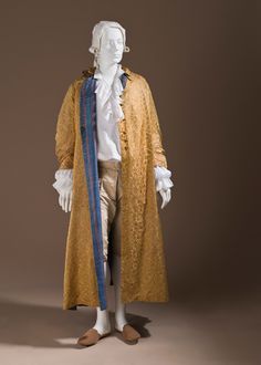 a fulllength image of a mannequin dressed in an 18thcentury period costume, set against a neutral brown backdrop. the mannequin has a white wig and wears a long, golden coat with a subtle patterned texture. the coat has a blue lining that is partially visible. underneath the coat, the mannequin wears a white shirt with ruffled lace cuffs. the lower body is clad in breeches and brown slippers. the mannequin is positioned in a standing pose.