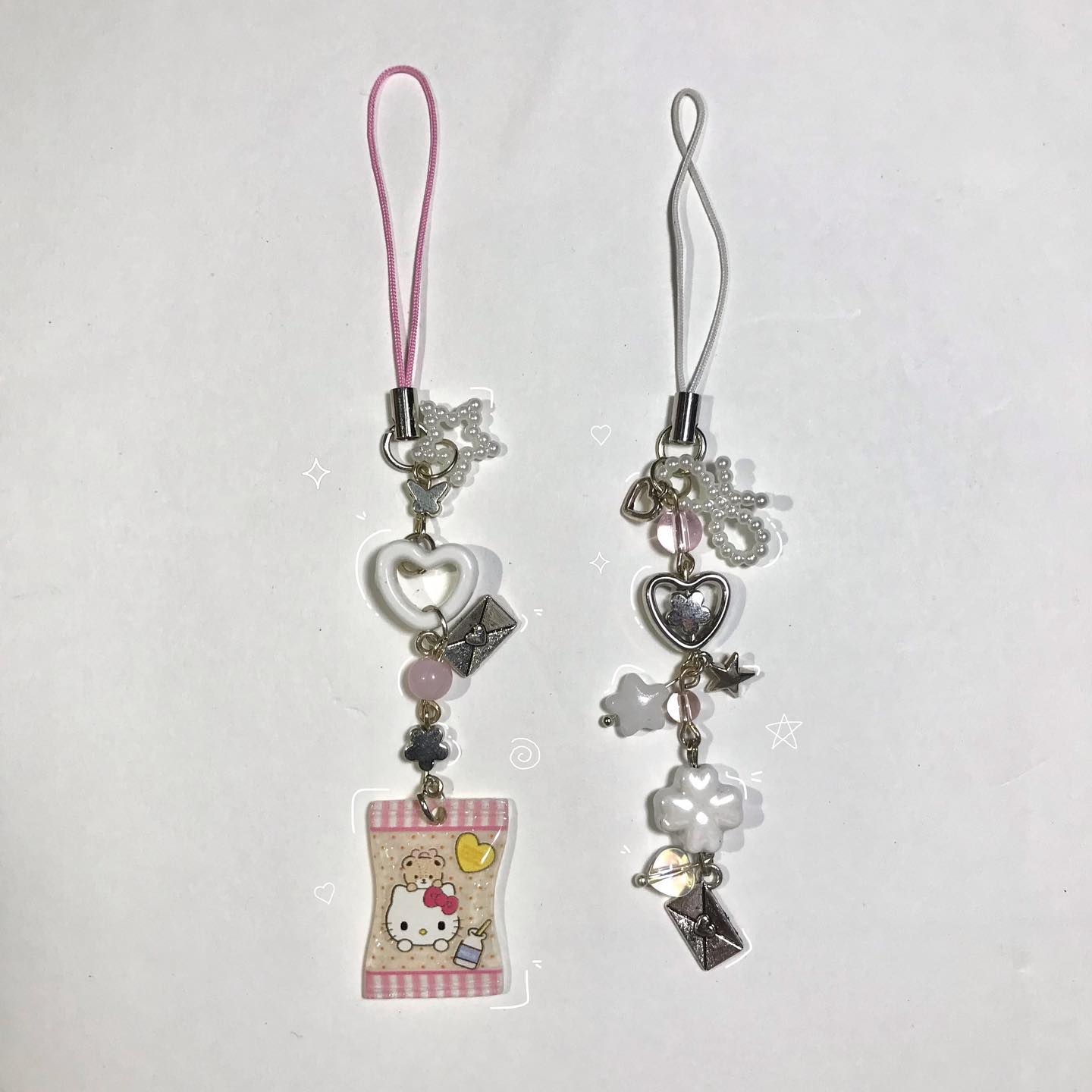 two phone charms are displayed against a white background. the charm on the left has a pink strap and features a white heart, a pink bead, and a hello kitty themed charm. the charm on the right has a white strap and features a silver heart, a white flower bead, and a silver letter charm. both charms have star and letter charms, as well as white and silver beads near the straps.