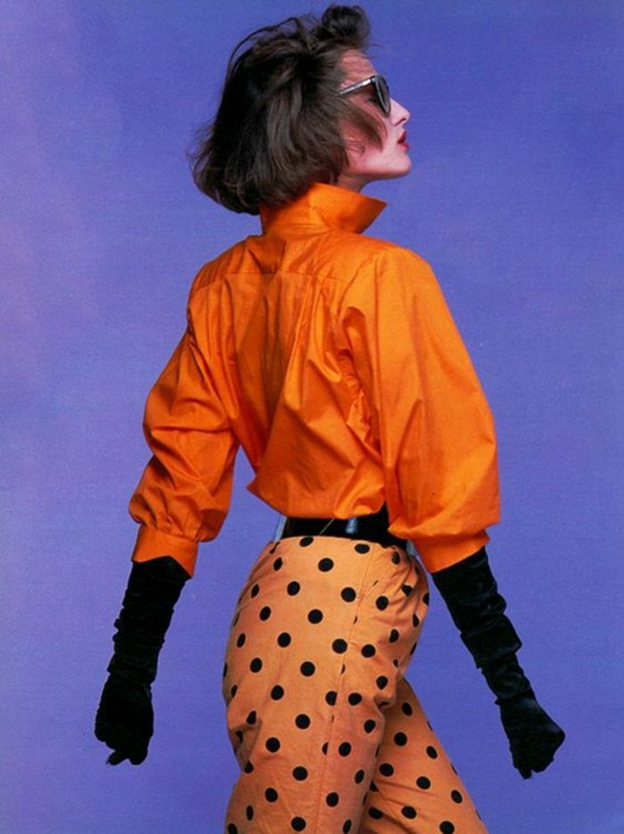 a woman poses in a vintagestyle fashion ensemble. she is wearing an orange highcollar shirt, polkadot pants, and long black gloves, complemented by sunglasses. the image features a studio shot with a purple background.