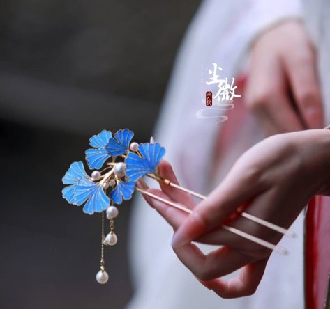 a woman wearing a white hanfu dress is holding a delicate blue ginkgo leaf hairpin adorned with pearls. the hairpin is crafted with intricate details, and the soft lighting highlights the beauty of the piece and the woman's hand.