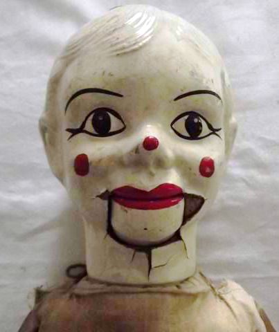 a close up of the face of a creepy vintage porcelain doll with a painted clown face. the doll has painted on red cheeks and a red smile. its eyes are black and white.  the doll is cracked around the eyes and mouth and has a faded look.