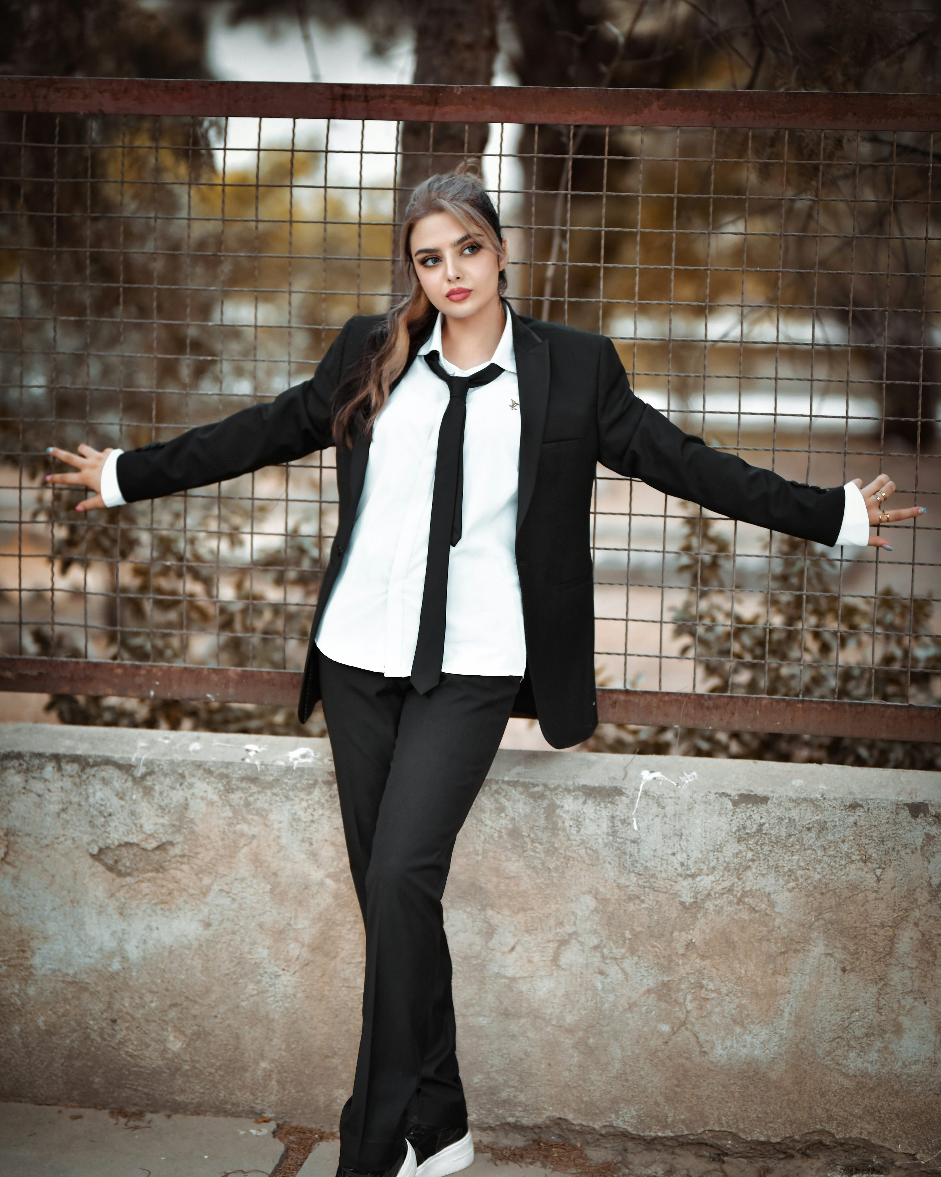 a young woman in a black pantsuit and white shirt with a black tie leans against a rusty metal fence. she is posing for a photo, her expression is serious, and she's looking off to her side. the background is an urban setting with a concrete wall and outoffocus trees. the overall style is fashionable and elegant. the image has a slightly moody or autumnal tone.