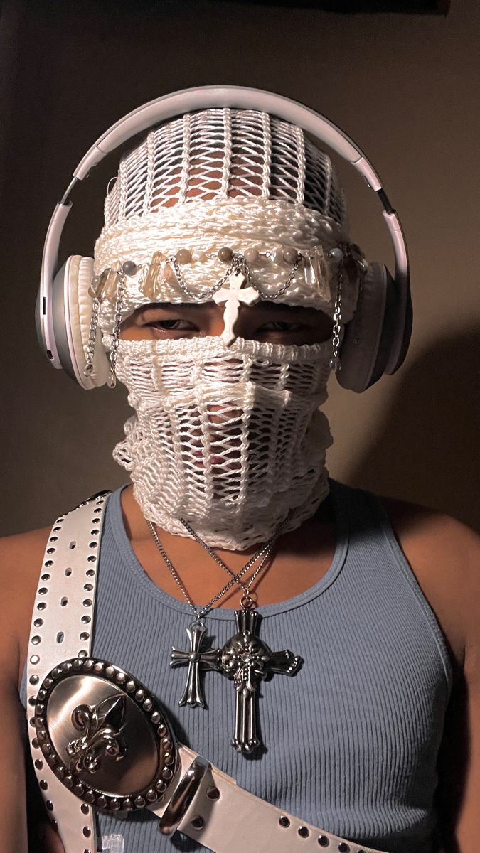 a person wearing a white facemask and headphones. they have two silver cross necklaces and a white belt with a silver buckle. they are wearing a blue tank top.  the person is looking directly at the camera.