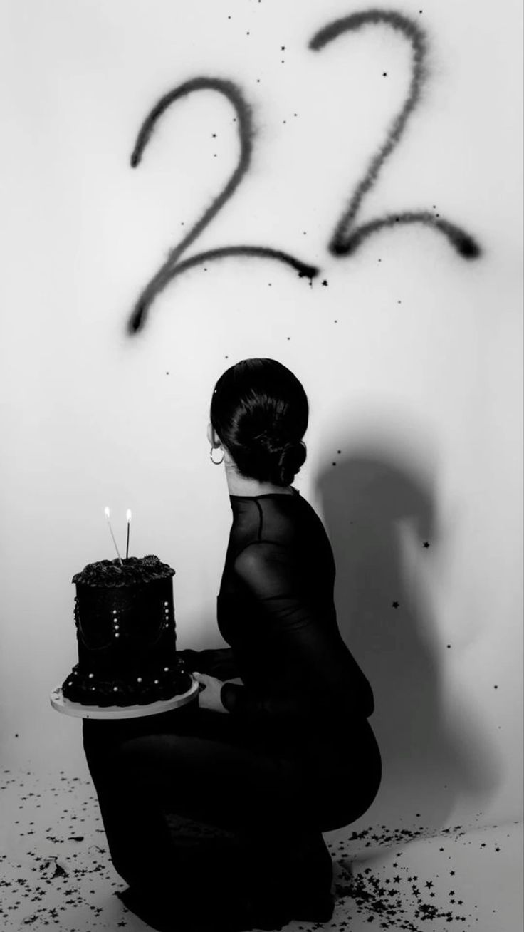 a woman in a black dress holds a black cake in front of her. she is celebrating her 22nd birthday. there are 22s written on the wall behind her and confetti on the floor.