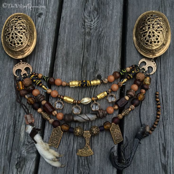 a necklace with multiple strands of different beads and bronze pendants is displayed on a weathered wooden surface. the necklace features oval brooches attached to the top, various types of beads in brown, gold, and orange tones, and pendants like a miniature thor's hammer and animal claws.