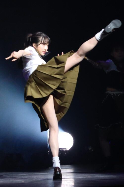 a young woman in a white shirt and green skirt is performing a high kick on a stage. she's wearing white socks and black shoes. the background is dark with a spotlight on her and another person standing in the shadow.
