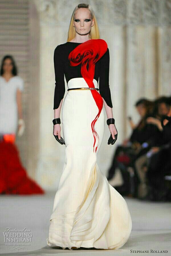 a beautiful fashion model in a black and white dress with a red accent walks down the runway. the dress is very elegant and sophisticated, with a flowing skirt and a fitted bodice. the model's hair and makeup are also very stylish, and she is posing confidently for the camera. the background is a blur of other models and photographers, but the model is the main focus of the image.