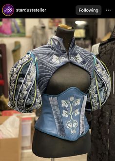 a closeup image showcasing a stunning handcrafted blue velvet corset with elaborate floral embroidery and matching detached sleeves. the corset, featuring a sweetheart neckline, is displayed on a dress form inside a studio.  the intricate detailing and craftsmanship suggest a piece inspired by historical or fantasy aesthetics.