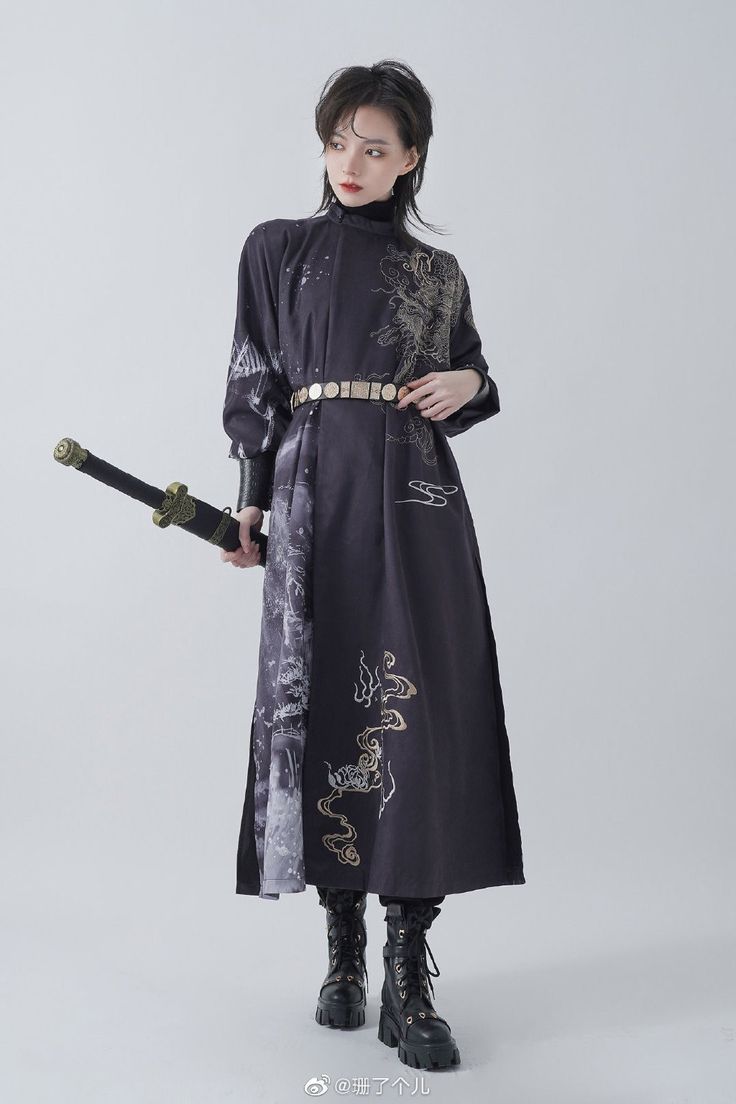a woman is posing in a studio against a white background.  she is wearing a long black dress with gold and white designs. she is wearing a gold belt and black boots.  she is holding a sword with a gold handle in her right hand.