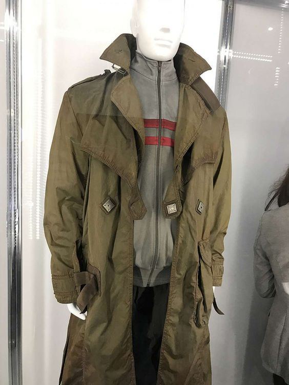 loseup view of an olive green long coat displayed on a mannequin. the coat is a trench coat style with several pockets, buttons, and a belted waist. underneath the coat is a grey zipup jacket with red stripes.  the mannequin is in a display case with a portion of a wall visible in the background.  a blurry portion of another person or object is visible in the lower right corner. the overall setting suggests a retail store or museum display.
