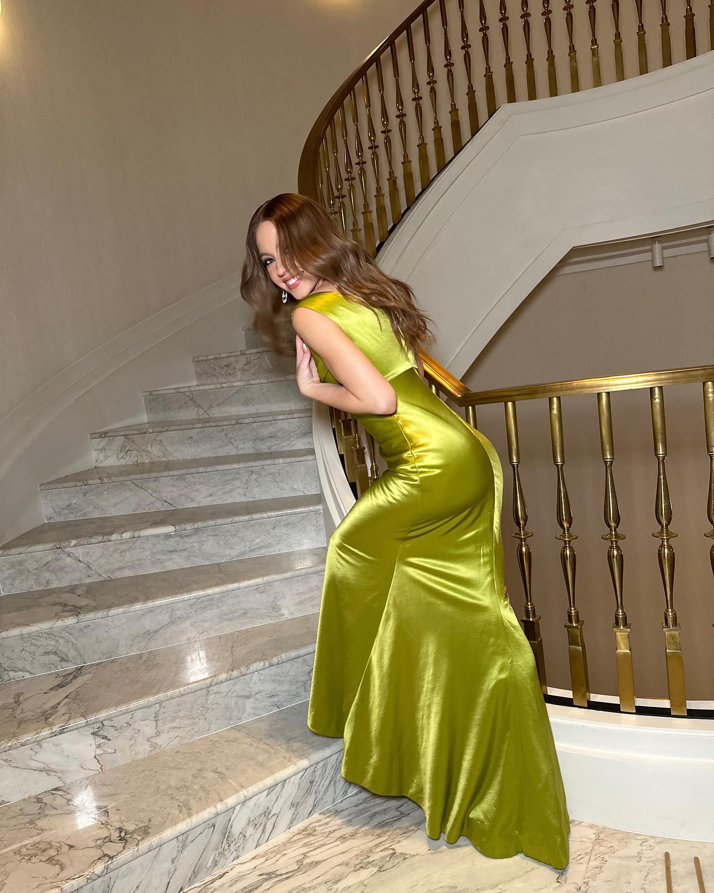 a young woman with long, wavy brown hair is posing on a marble staircase in a shimmering lime green satin gown.  she's turned slightly away from the camera, looking back over her shoulder with a slight smile. the dress is formfitting and has a mermaid or fishtail style. the staircase is grand and features a gold railing. the overall ambiance is luxurious and elegant.
