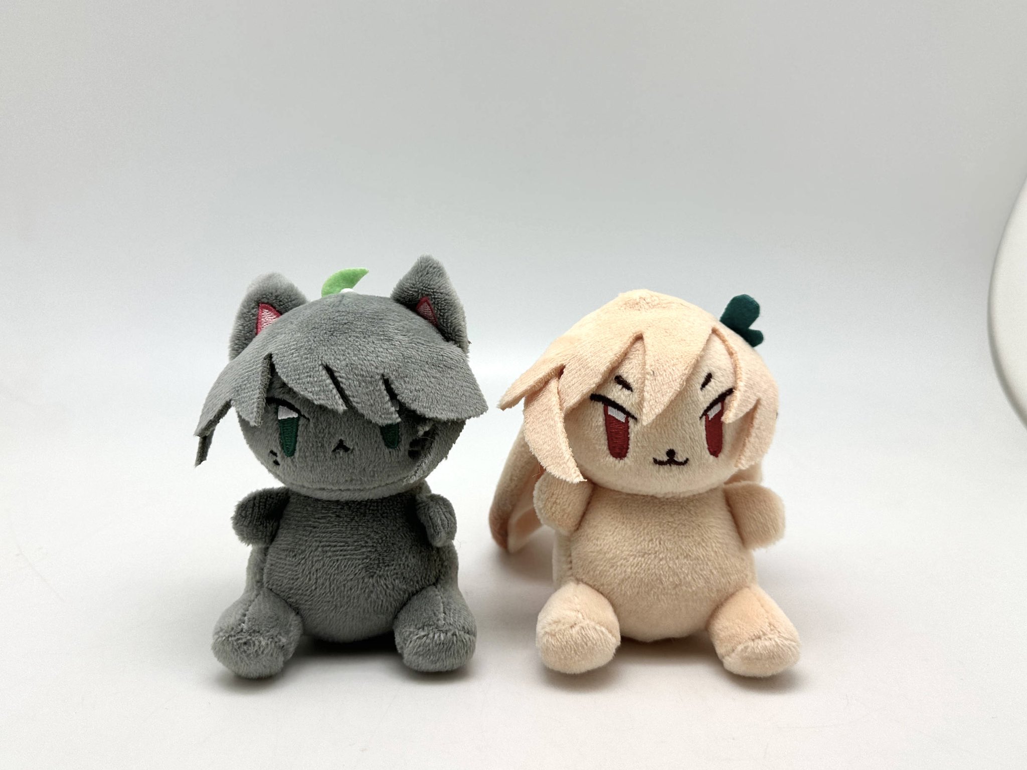 two cute plush toys, a gray cat and a white bunny with pink ears, sitting sidebyside on a white surface.  the cat has green eyes, a green leaf on its head, and a serious expression. the bunny has red eyes, a green flower on its head, and a friendly smile.