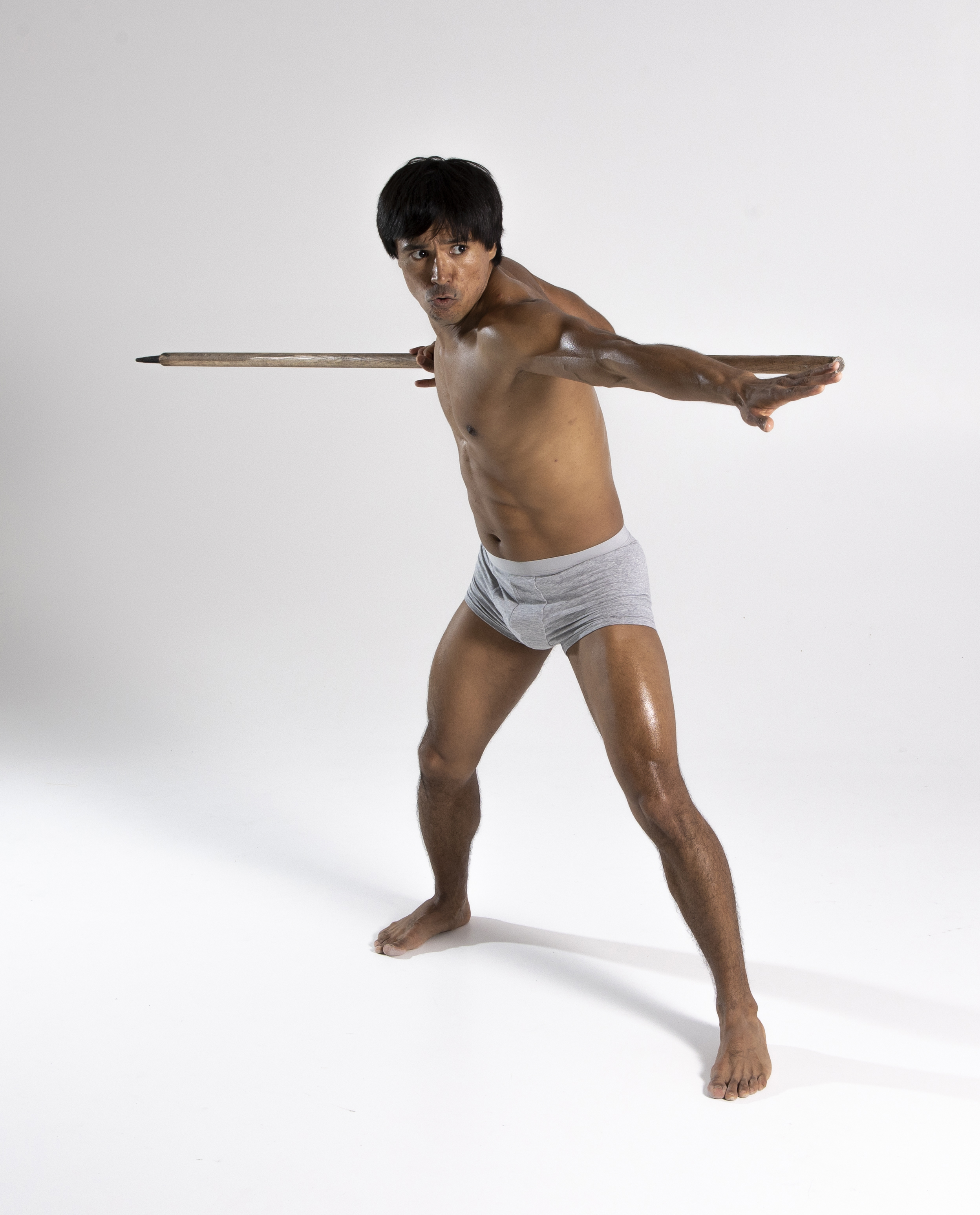 a shirtless man in gray underwear is posed in a martial arts stance, wielding a wooden pole. he has a muscular build and a determined expression. the photo is a studio shot with a plain white background.