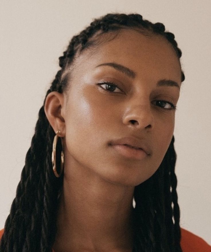 closeup portrait of a young black woman with long, thick braids. she's wearing gold hoop earrings and has a serious, neutral expression. her skin is beautifully lit and appears healthy and glowing. the background is a simple, light beige. the overall style is fashion and beauty oriented, with a focus on the model's face and hair.