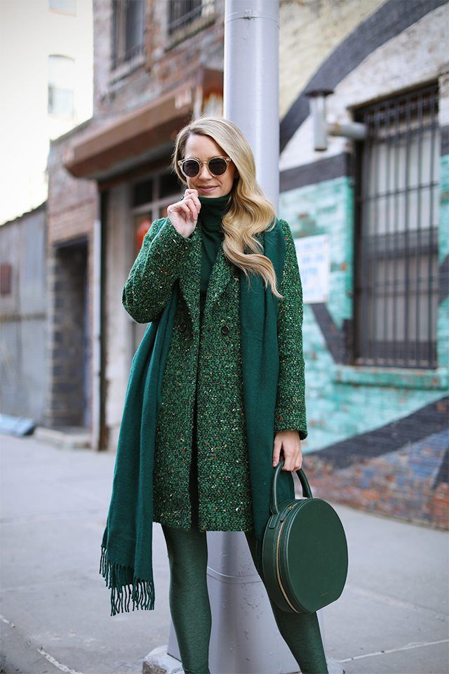 the image showcases a fashionably dressed blonde woman standing on a city street. she's wearing a kneelength, emerald green tweed coat over a matching turtleneck sweater and green tights.  a round, dark green handbag hangs from her arm, and she accessorizes with sunglasses. the overall look is monochromatic and stylish, embodying a winter or fall street style aesthetic. the background features an urban setting with a brick building and a tealpainted wall.