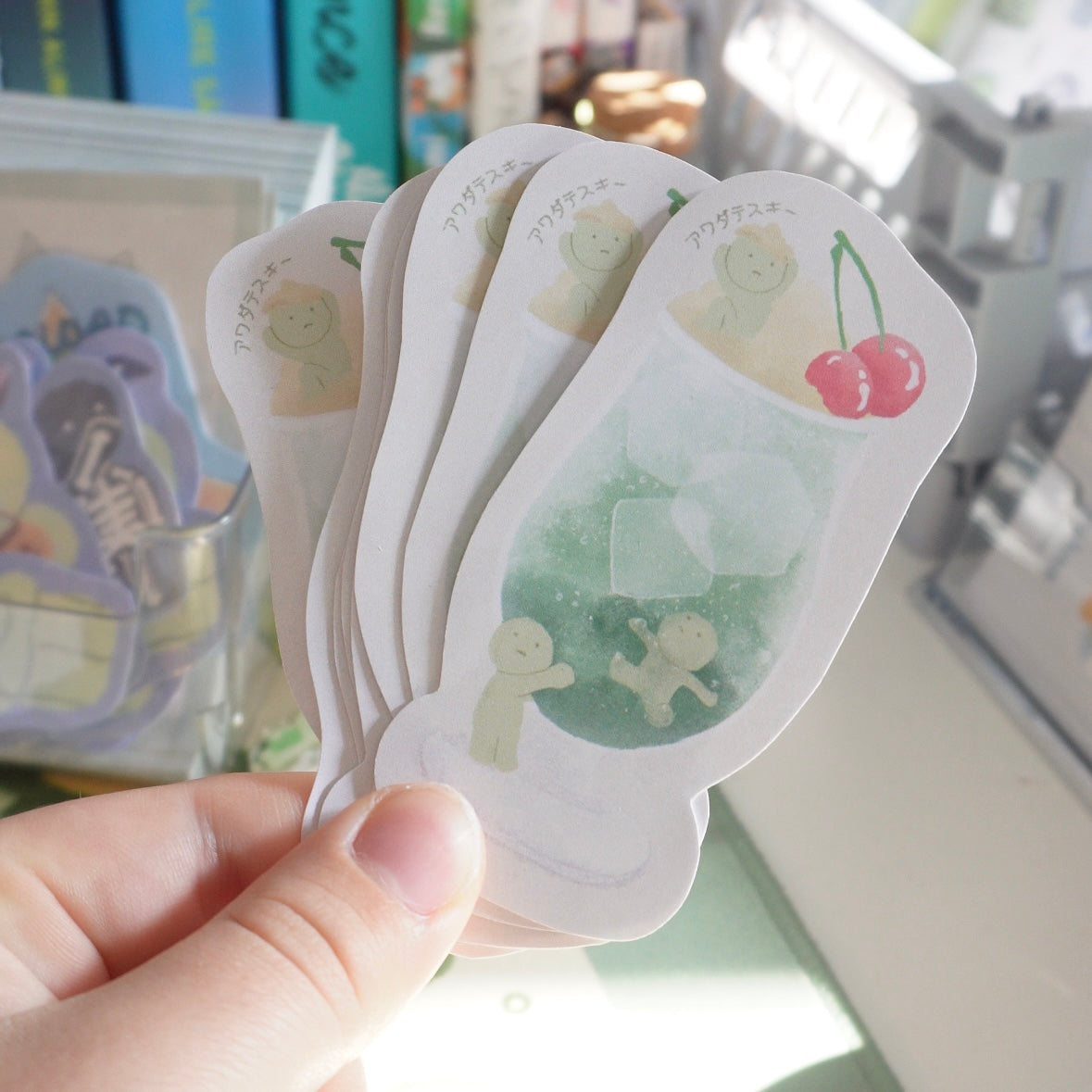 a hand is holding a stack of cute, pastelcolored japanese stickers. the stickers feature cartoon characters and desserts. the top sticker shows a drink with cherries on top. the stickers are all white with pink and green accents.