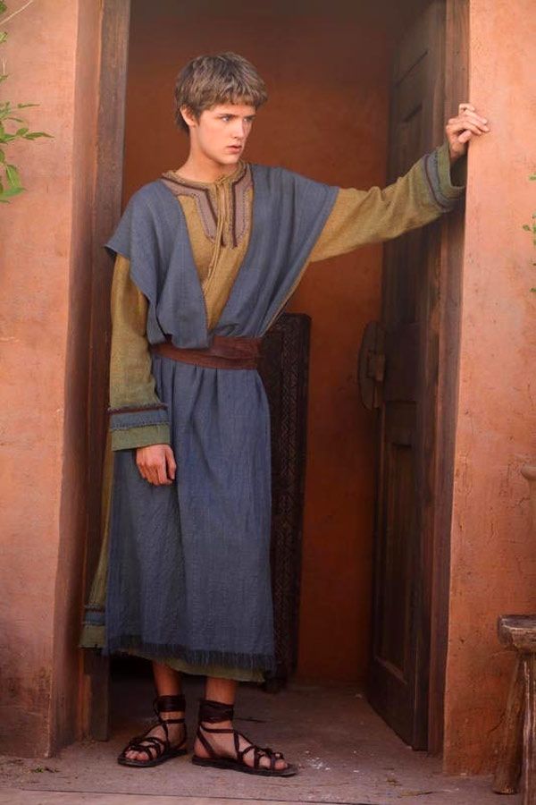a young man with fair hair stands in a doorway, wearing a long blue robe cinched at the waist with a brown belt. underneath the robe, he has on a tan tunic with long sleeves. his footwear consists of dark brown leather sandals that lace up around his ankles. he is leaning slightly to his right, with his right hand resting on the door frame. the walls surrounding the doorway are a warm, earthy reddishbrown color. a simple wooden stool is visible in the bottom right corner of the image.