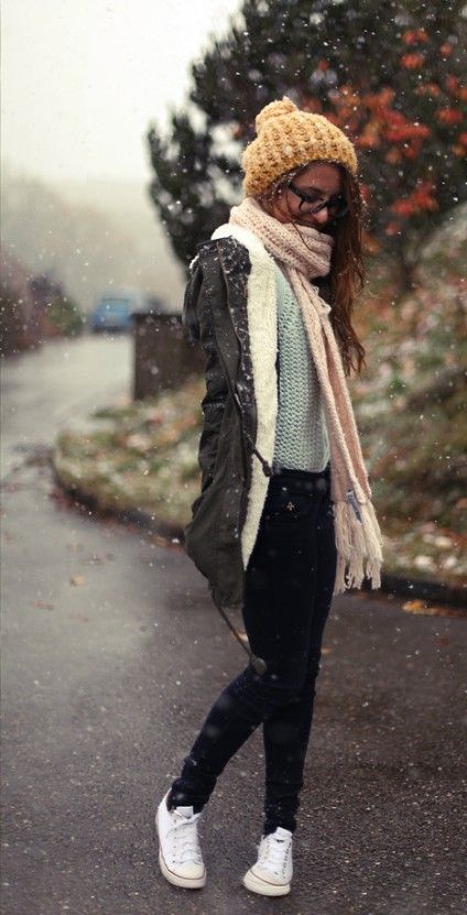 a young woman walking down a snowy street, wearing a warm winter coat, beanie, scarf, and jeans. she is looking down at the ground and smiling. the weather is cold, and it is snowing.