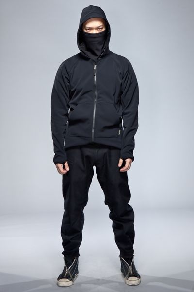 a man is standing in front of a grey background, wearing a black techwear outfit. he is wearing a black hoodie with a builtin face mask, black pants, and black and white sneakers. he is posing with his arms at his sides and his feet slightly apart.