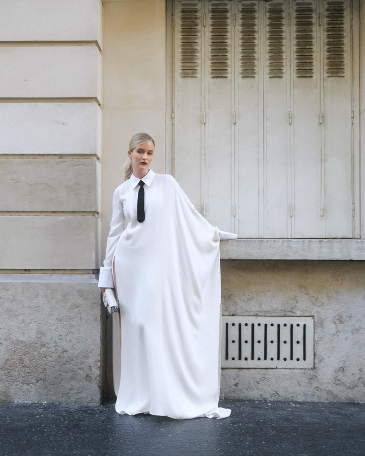 a woman wearing a long, white dress with a black tie stands in front of a building. she is also wearing white gloves and has a blonde ponytail. the building is a light color and has a large, closed window behind her.