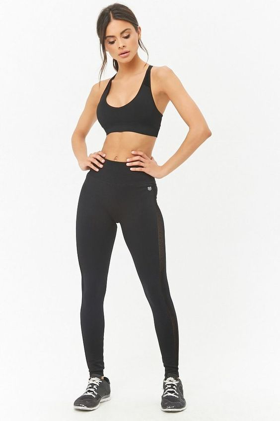 a woman is modeling a black activewear set against a white background. she is wearing a black sports bra and black leggings with mesh paneling. she is standing with her hands on her hips and her feet slightly apart.