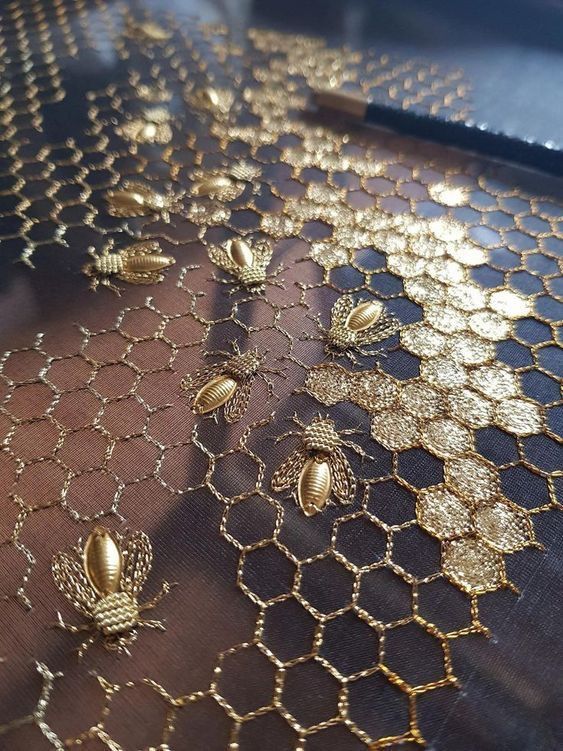 a closeup shot of fabric embroidered with golden honeycomb and bee designs. the honeycomb pattern is intricately stitched, and the bees are scattered across the fabric.