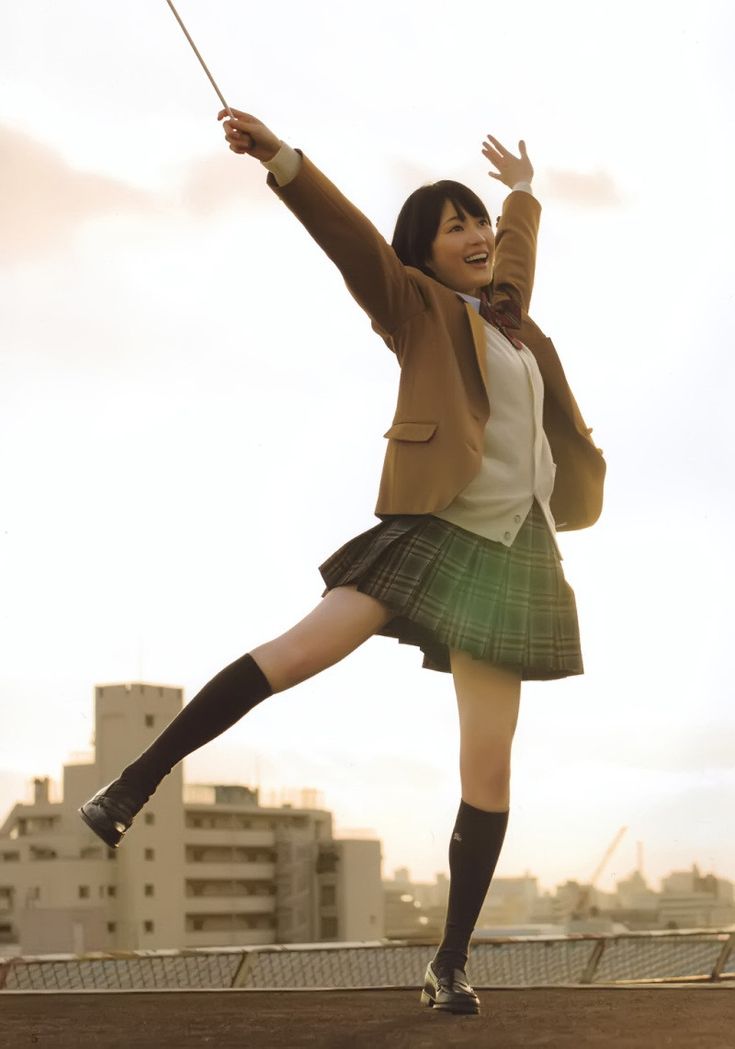 a young japanese woman in a school uniform is standing on a rooftop with her arms spread wide and a big smile on her face. she is wearing a brown jacket, white shirt, black skirt, black socks, and black shoes. the setting sun is casting a warm glow on her and the city skyline behind her.